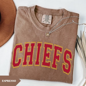 May include: A brown short-sleeved t-shirt with a vintage-style wash, featuring the word CHIEFS in large red block letters with a yellow outline. The shirt has a Comfort Colors brand label at the neck and is paired with a silver-toned chain necklace.