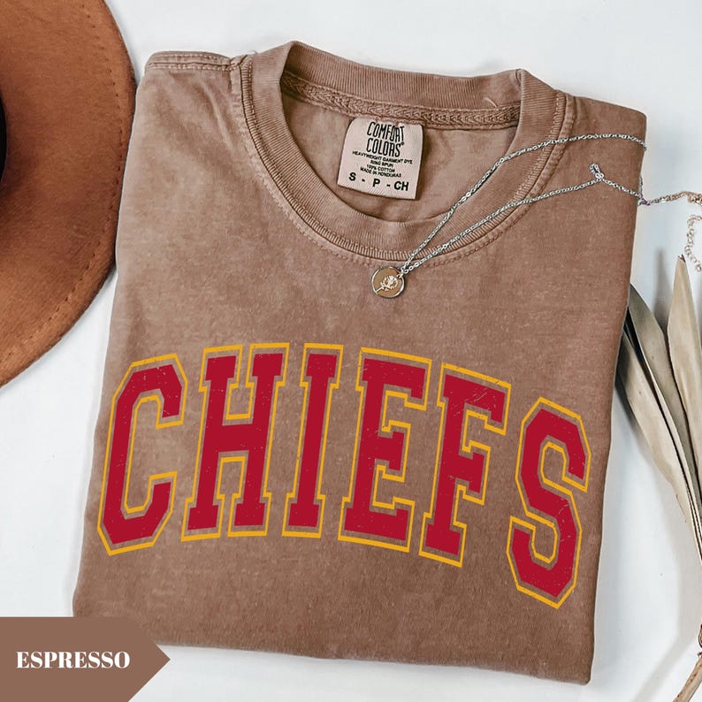 May include: A brown short-sleeved t-shirt with a vintage-style wash, featuring the word CHIEFS in large red block letters with a yellow outline. The shirt has a Comfort Colors brand label at the neck and is paired with a silver-toned chain necklace.