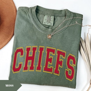 May include: A moss green short-sleeve t-shirt featuring the word CHIEFS printed in large red block letters with a yellow outline. The shirt has a crew neckline and a Comfort Colors brand label on the inside collar indicating a size small.