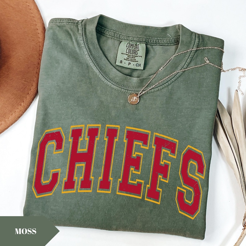 May include: A moss green short-sleeve t-shirt featuring the word CHIEFS printed in large red block letters with a yellow outline. The shirt has a crew neckline and a Comfort Colors brand label on the inside collar indicating a size small.