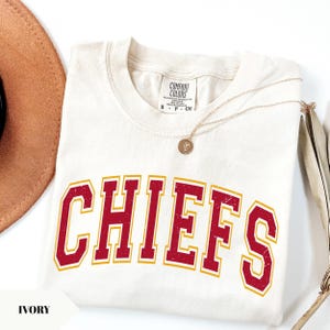 May include: An ivory t-shirt featuring the word CHIEFS in a distressed red font with a gold outline. The shirt has a crew neckline and a Comfort Colors brand label at the neck. A thin gold chain necklace with a small circular pendant rests on the shirt.