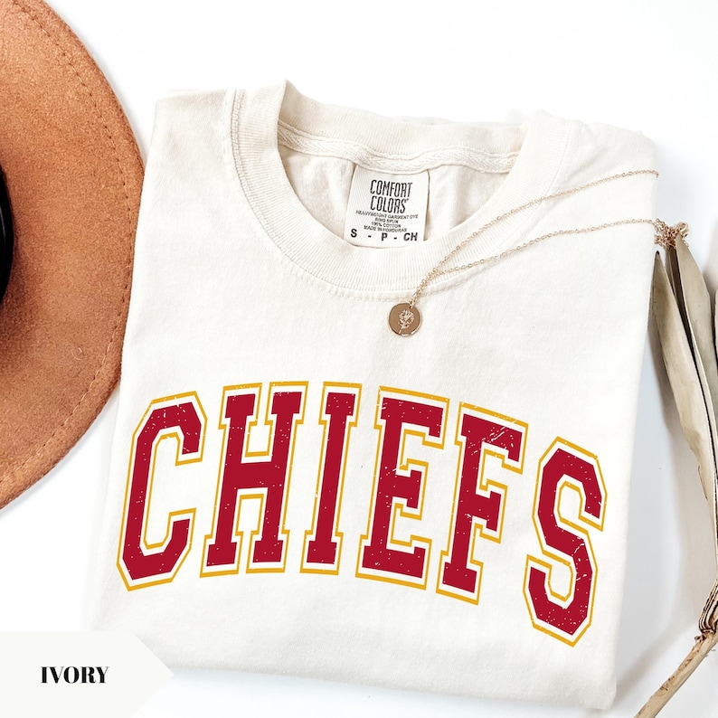 May include: An ivory t-shirt featuring the word CHIEFS in a distressed red font with a gold outline. The shirt has a crew neckline and a Comfort Colors brand label at the neck. A thin gold chain necklace with a small circular pendant rests on the shirt.