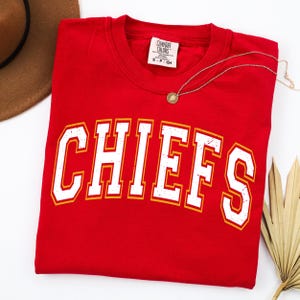 May include: A red short-sleeved t-shirt featuring the word CHIEFS in large, white, distressed-style block letters with a yellow outline. The shirt has a Comfort Colors brand label at the inner neckline and is displayed with a gold-toned necklace.