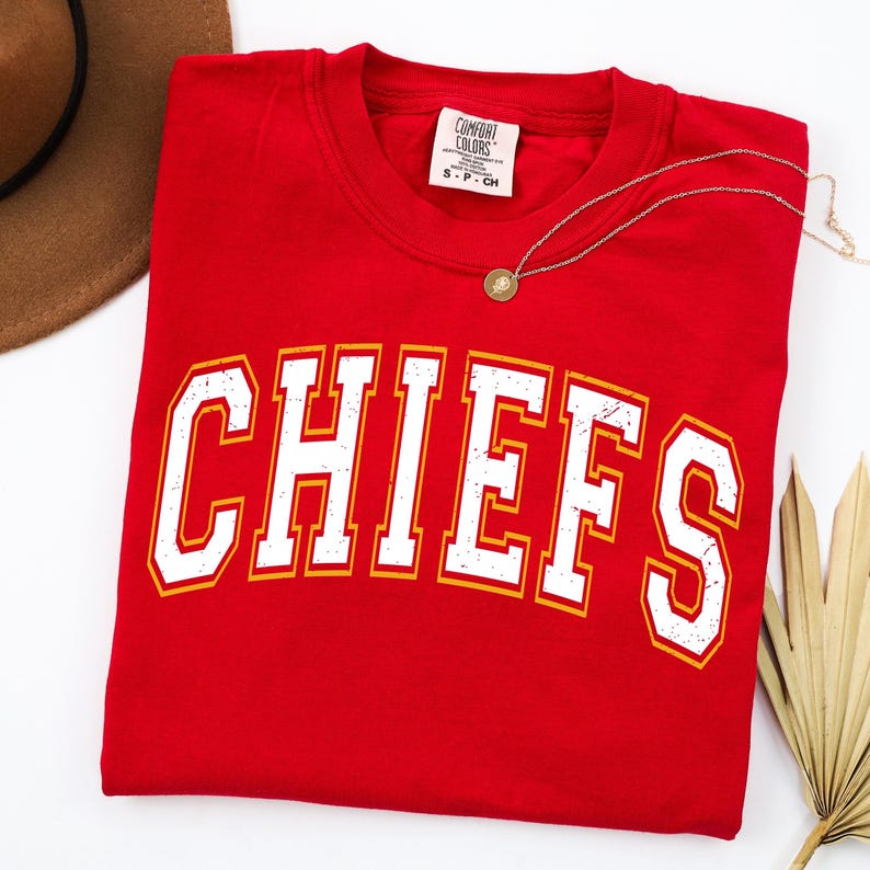 May include: A red short-sleeved t-shirt featuring the word CHIEFS in large, white, distressed-style block letters with a yellow outline. The shirt has a Comfort Colors brand label at the inner neckline and is displayed with a gold-toned necklace.