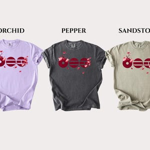 May include: Three short-sleeve crew neck t-shirts in orchid, pepper, and sandstone colors. Each shirt features a graphic of three dark red circles with horizontal lines and pink cherry blossom branches extending from the design across the chest.