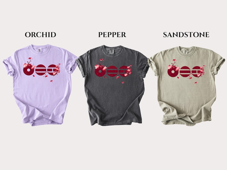 May include: Three short-sleeve crew neck t-shirts in orchid, pepper, and sandstone colors. Each shirt features a graphic of three dark red circles with horizontal lines and pink cherry blossom branches extending from the design across the chest.
