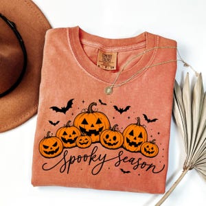Könnte beinhalten: Ein rostorangefarbenes T-Shirt mit einem schwarzen Aufdruck von sechs verschieden großen Kürbissen, mehreren fliegenden Fledermäusen und dem Schriftzug "Spooky season" in Schreibschrift. Das T-Shirt hat einen Rundhalsausschnitt und ein Comfort Colors Etikett.