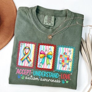 Könnte beinhalten: Olivgrünes Comfort Colors T-Shirt mit farbenfrohem Autismus-Awareness-Design. Das Design umfasst ein Regenbogenband, eine Schleife und ein Puzzleteil, mit den Worten "ACCEPT UNDERSTAND LOVE, autism awareness."
