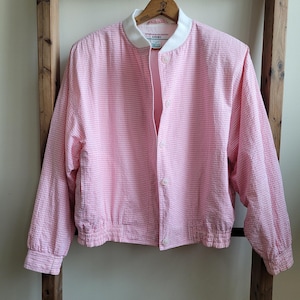 May include: A pink and white checkered bomber jacket with a white collar and button closure. The jacket is hanging on a wooden hanger. The jacket is made of a textured fabric.