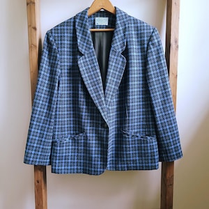 May include: A blue and black houndstooth blazer with a notched collar and two front pockets. The jacket is hanging on a wooden hanger. The blazer has a classic, tailored fit.