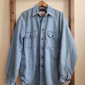 May include: A light blue denim long-sleeve shirt featuring a button-down front, a pointed collar, and two chest pockets with button closures. The interior is lined with a dark brown and black plaid fabric, and the inner neck label reads American Eagle.