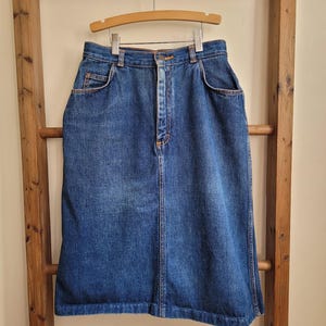 May include: A high-waisted blue denim skirt featuring a classic five-pocket design, a front button and zipper closure, belt loops, and visible orange stitching. The skirt has a straight silhouette that falls below the knee with a hemmed bottom edge.