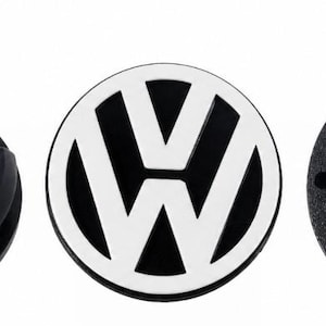 May include: Three circular wheel centre caps. The left cap is black with four mounting clips. The centre cap features a black and white Volkswagen logo. The right cap is a solid black textured disc with four small circular indentations.
