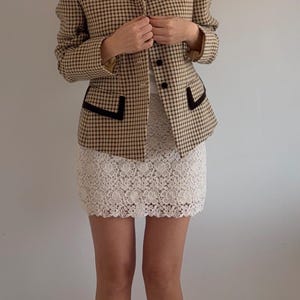 May include: A tan and black checkered blazer with black trim and a white lace skirt. The blazer has a classic collar and button closure. The skirt is a mini skirt with a floral lace pattern. The outfit is a vintage style.