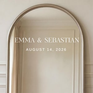 May include: A large, arched mirror with a silver frame. The mirror reflects a light-colored room. The text "EMMA & SEBASTIAN" and "AUGUST 14, 2026" are displayed in white, centered on the mirror's surface. The overall aesthetic is elegant and minimalist.