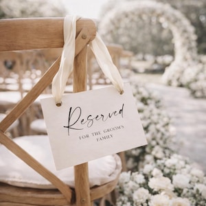 Könnte beinhalten: Ein Holzstuhl mit einem cremefarbenen Schild mit der Aufschrift "Reserved FOR THE GROOM'S FAMILY". Das Schild ist mit cremefarbenen Bändern am Stuhl befestigt. Im Hintergrund ein Blumenbogen und weitere Stühle.