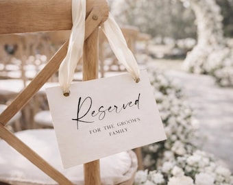 May include: A wooden chair with a cream-colored sign that reads "Reserved FOR THE GROOM'S FAMILY". The sign is attached to the chair with cream-colored ribbons. The background features a floral arch and other chairs.