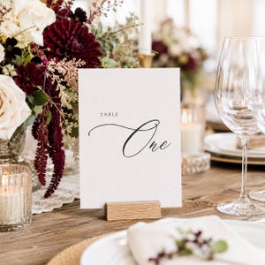 May include: A white table card with the word "Table" above and the word "One" in elegant script. The card is held by a small wooden block, set on a wooden table. The background includes floral arrangements, candles, and glassware.