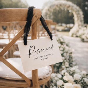 May include: A cream-coloured sign reading "Reserved" in black script, tied to a wooden chair with a black ribbon. The text below the main word reads "For the Groom's Family". The scene suggests a wedding.