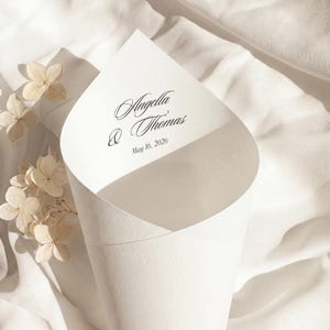 Könnte beinhalten: Ein weißer Papierkegel mit den Namen "Angella & Thomas" und dem Datum "16. Mai 2026" in schwarzer Schrift. Der Kegel befindet sich auf einem weißen Stoffhintergrund mit getrockneten Blumen.