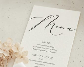 Może przedstawiać: Kremowa karta menu ze słowem "Menu" w eleganckim czarnym skrypcie. Menu zawiera opcje sałatek i przystawek, w tym sałatkę z kraba i języka wołowego. Suszone kwiaty i biała wstążka na pierwszym planie.