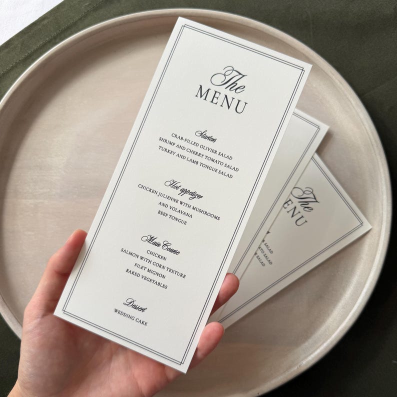 May include: A tall, rectangular white paper menu with black text and a double-line border. The menu lists sections for Starters, Hot appetizer, Main Course, and Dessert, including items like Crab-filled Olivier salad, Chicken, and Wedding Cake.