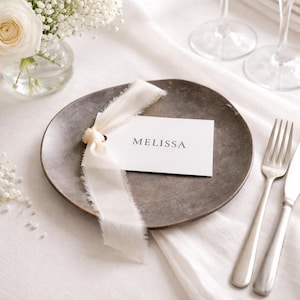 May include: A table setting featuring a grey plate, cutlery, and a white card with the name "MELISSA". The card is tied with a cream ribbon. A small vase of white flowers is visible in the background.