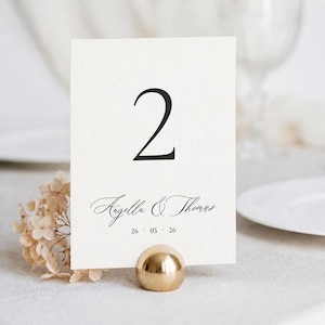 May include: A cream-colored table number card with the number 2 in black script. The card features the names Angella & Thomas and the date 26-05-26. The card is held by a gold-colored metal sphere.