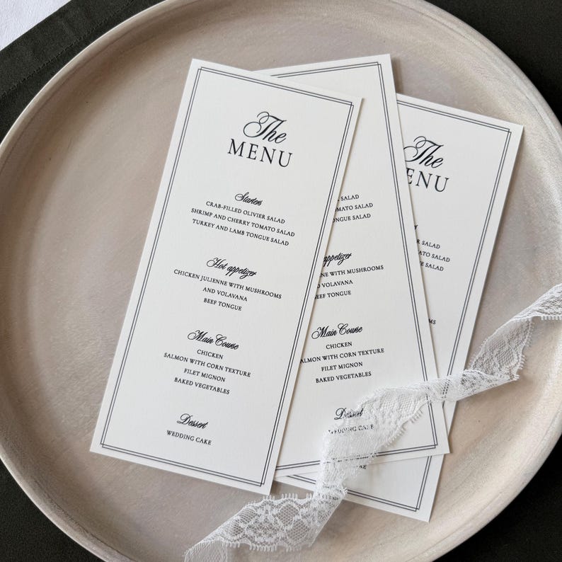 May include: Three tall, rectangular white paper menus with black text and a double-line border. The menus feature headings for Starters, Hot appetizer, Main Course, and Dessert, with a delicate white lace ribbon draped across the bottom of the cards.