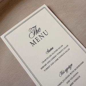May include: A rectangular off-white menu card featuring a double-line black border. The text reads The Menu, Starters, Crab-filled Olivier Salad, Shrimp and Cherry Tomato Salad, Turkey and Lamb Tongue Salad, Hot appetizer, Chicken Julienne with Mushrooms and Volavana, and Beef Tongue.