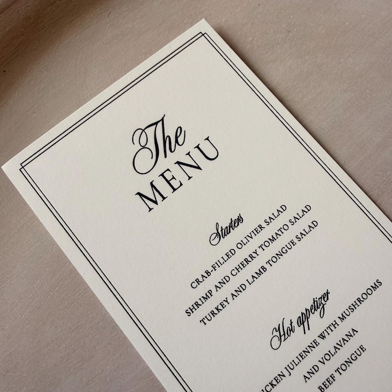May include: A rectangular off-white menu card featuring a double-line black border. The text reads The Menu, Starters, Crab-filled Olivier Salad, Shrimp and Cherry Tomato Salad, Turkey and Lamb Tongue Salad, Hot appetizer, Chicken Julienne with Mushrooms and Volavana, and Beef Tongue.