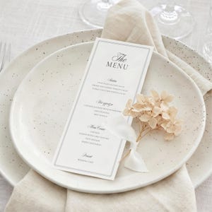 May include: A white rectangular menu card with black text titled The Menu, featuring sections for Starters, Hot appetizer, Main Course, and Dessert. The card is placed on a speckled white plate with a small bouquet of dried cream flowers and a white ribbon.