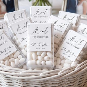 May include: A collection of clear rectangular plastic containers filled with white mints. Each container features a white label with black text reading Mint FOR EACH OTHER, Ethan & Olivia, and the date 09 / 16 / 2026, arranged in a woven basket.