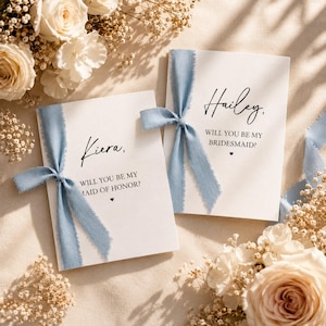 Può includere: Due cartoncini rettangolari bianchi con testo nero e fiocchi di nastro blu polvere sfilacciato. Un cartoncino riporta Kiera, WILL YOU BE MY MAID OF HONOR? e l'altro Hailey, WILL YOU BE MY BRIDESMAID? Ogni cartoncino ha un piccolo cuore nero.
