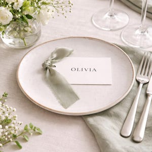Puede incluir: Un plato de cerámica blanca moteada con una tarjeta con el nombre "OLIVIA" atada con una cinta verde salvia. Cubiertos de plata y una servilleta de lino verde están colocados a la derecha. Un jarrón de flores blancas está en el fondo.