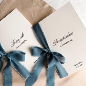 Puede incluir: Dos tarjetas de boda color marfil con lazos de cinta color turquesa. Las tarjetas dicen "A mi esposa" y "A mi esposo" con la fecha "26-05-2026" impresa. Las tarjetas están atadas con una cinta color turquesa.