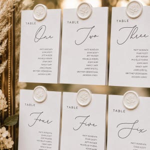 May include: Six white table cards with calligraphy numbers and guest names, topped with white wax seals with a floral design. The cards are arranged in a grid, suitable for weddings or events. The numbers are written in elegant calligraphy.