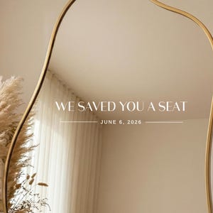 May include: A wall mirror with a thin gold frame and an irregular, wavy shape. The glass surface displays the white text WE SAVED YOU A SEAT and the date JUNE 6, 2026, centered between two horizontal lines, reflecting a room with curtains.