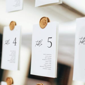 May include: White table cards with black text, held by gold wax seals. The cards are labeled with table numbers and guest names. The cards are hanging, creating a clean and elegant display.