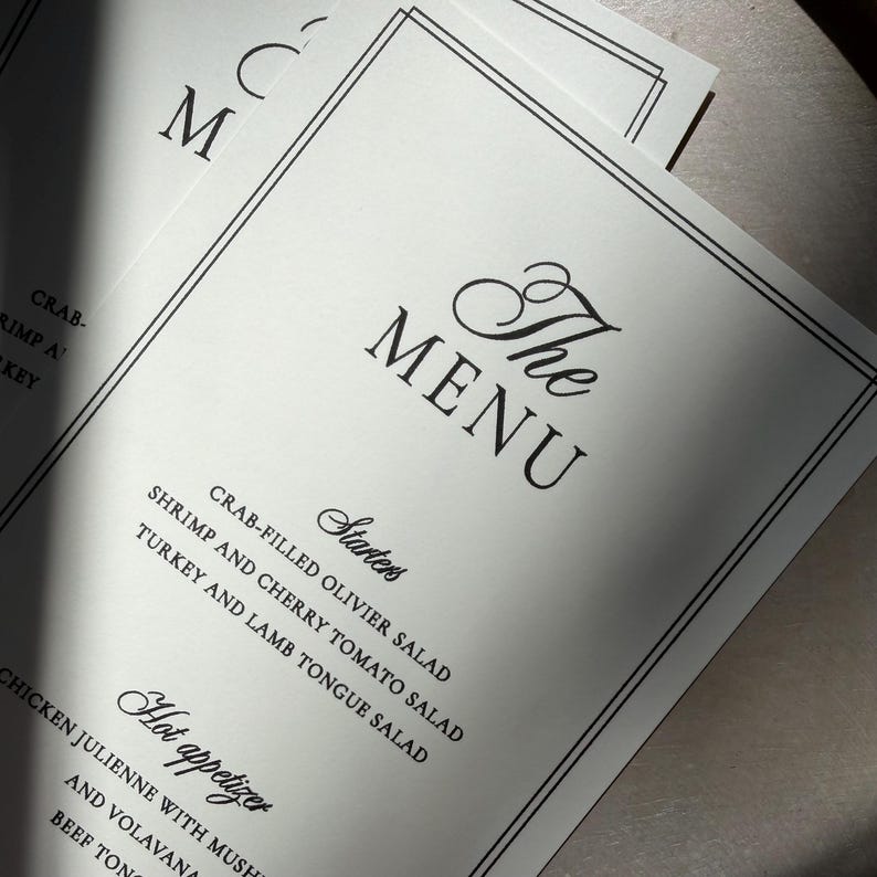 May include: A white paper menu with black text and a double-line border. The header reads The MENU, followed by Starters including Crab-filled Olivier salad, Shrimp and cherry tomato salad, and Turkey and lamb tongue salad, plus Hot appetizer.