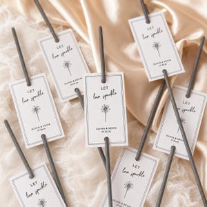 Könnte beinhalten: Mehrere graue Wunderkerzen, die jeweils an einem rechteckigen weißen Papieranhänger befestigt sind. Die Anhänger haben einen schwarzen Doppellinienrand, den Text "LET love sparkle", eine kleine Sternengrafik sowie die Namen "OLIVIA & KEVIN" mit dem Datum "07.25.26."