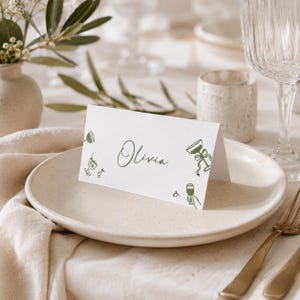 May include: A white folded place card featuring the name Olivia written in a dark green cursive script. The card is decorated with small, minimalist dark green line art illustrations of various cocktail glasses and a pair of cherries.