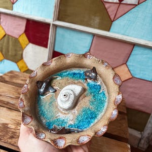 Op de afbeelding: Een handgemaakte keramische kom met een schelp- en aquatisch ontwerp. De kom heeft een geschulpte rand en een getextureerd, zanderig interieur met blauwe en turquoise accenten, en kleine, donkergekleurde zeedieren.