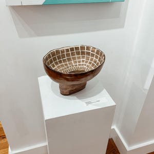 May include: A brown ceramic bowl with a grid pattern interior, displayed on a white pedestal. The bowl has a wide, open top and a rounded base. The pedestal is a simple, square shape. The background includes a painting with teal and white colors.