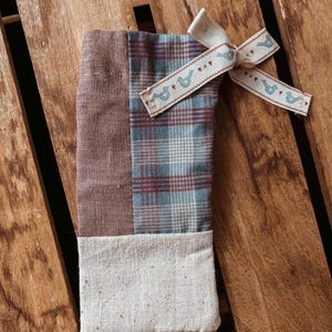 Puede incluir: Una bolsa de regalo de tela hecha a mano con un diseño de patchwork. La bolsa presenta secciones de tela marrón, a cuadros y blanquecina. Una cinta color crema con un estampado de pájaros está atada con un lazo. La bolsa está sobre una superficie de madera.