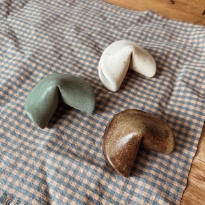 Può includere: Tre biscotti della fortuna in ceramica, verdi, crema e marroni, poggiano su un tessuto a quadretti blu e beige. I biscotti sono disposti a triangolo, mostrando la loro forma curva e le superfici strutturate.