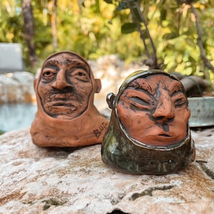 May include: Two ceramic sculptures of human faces. The sculpture on the left is a terracotta color with a mustache. The sculpture on the right has a dark green glaze around the face. Both sculptures are on a rock.