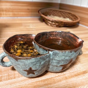 May include: A handmade ceramic mug with two connected bowls, glazed in a mottled blue-green with brown accents. Each bowl features a brown star. One bowl contains a herbal tea with visible chamomile flowers.