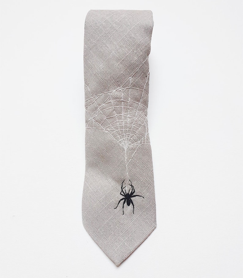 Eine graue Krawatte aus Leinen mit einem weißen gestickten Spinnennetz und einer schwarzen Spinne, die am Netz hängt.
