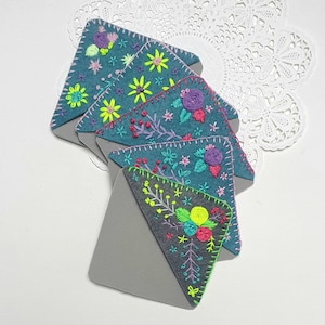 May include: Set of five teal blue felt bookmarks with colorful embroidered floral designs. Each bookmark has a gray felt backing.